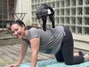Photo of someone smiling, doing Yoga with a small goat on their back.